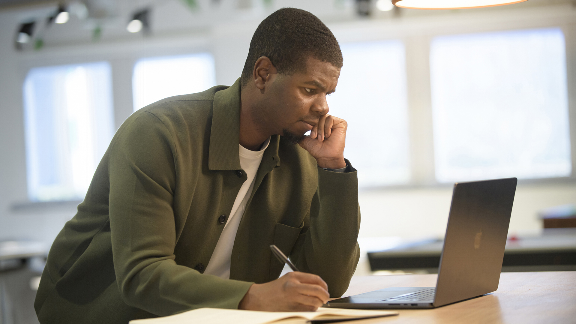 A man writing in a notebook while looking at a laptop screen.
