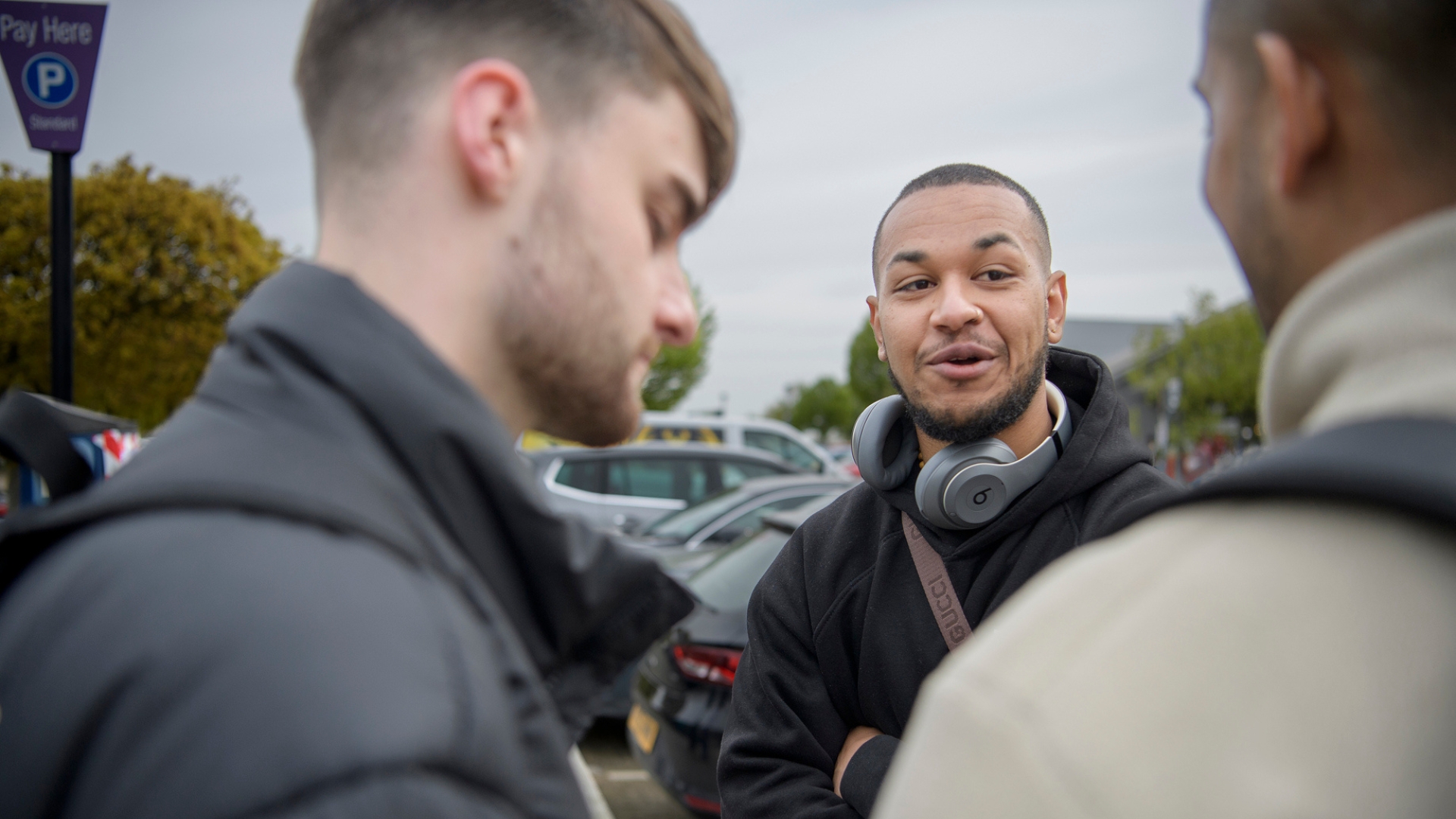 Three people talking together in a car park with parked cars behind them.