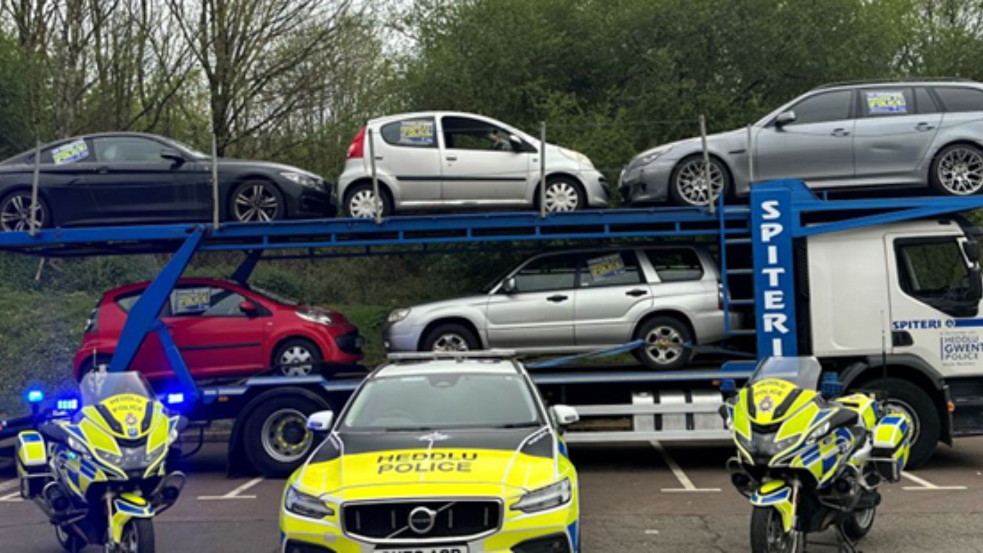 Two Police motorbikes surround a police car while a lorry carrying cars is in the background