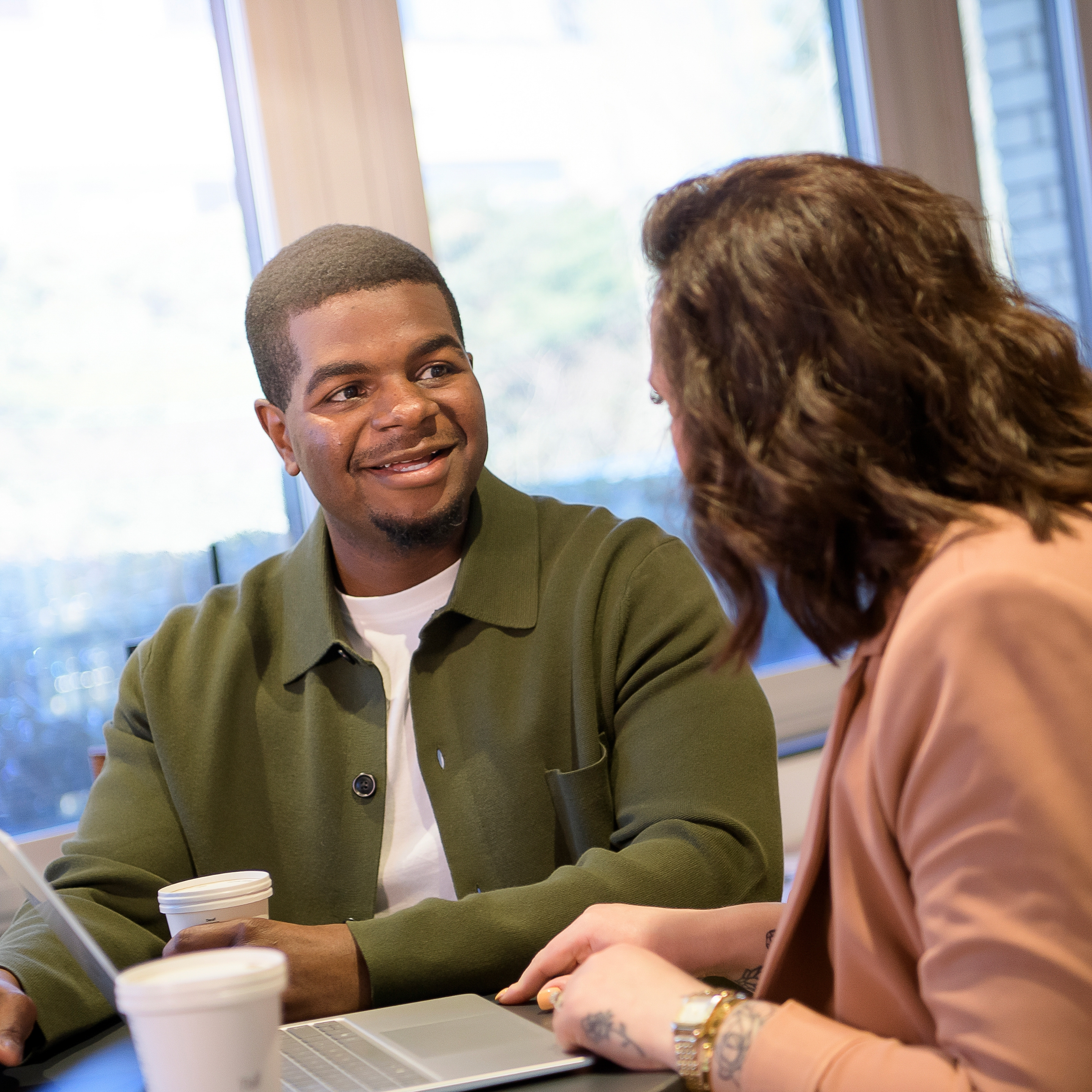 Two people sitting at a table talking, with a laptop and coffee cups.