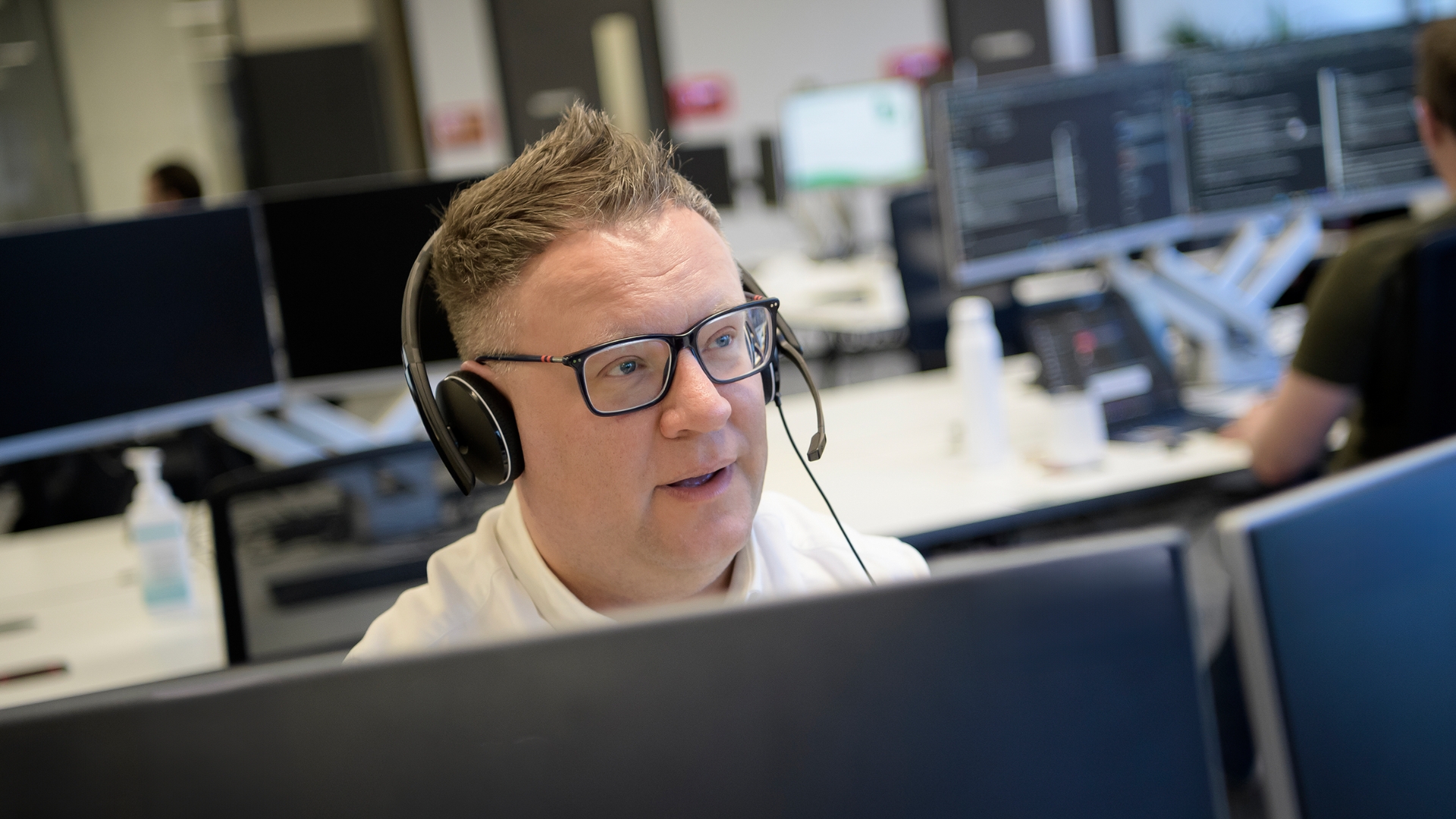 A man wearing a headset working at a computer, with other desks and screens in the background.