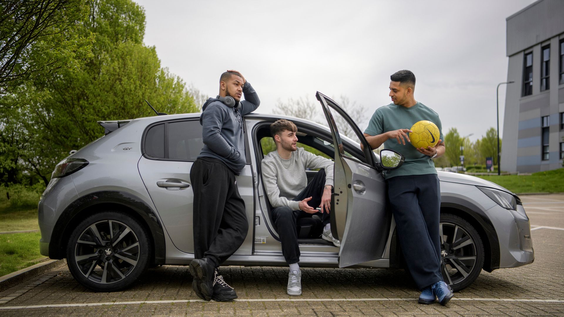 Three young men standing beside a parked car in a car park, one holding a football