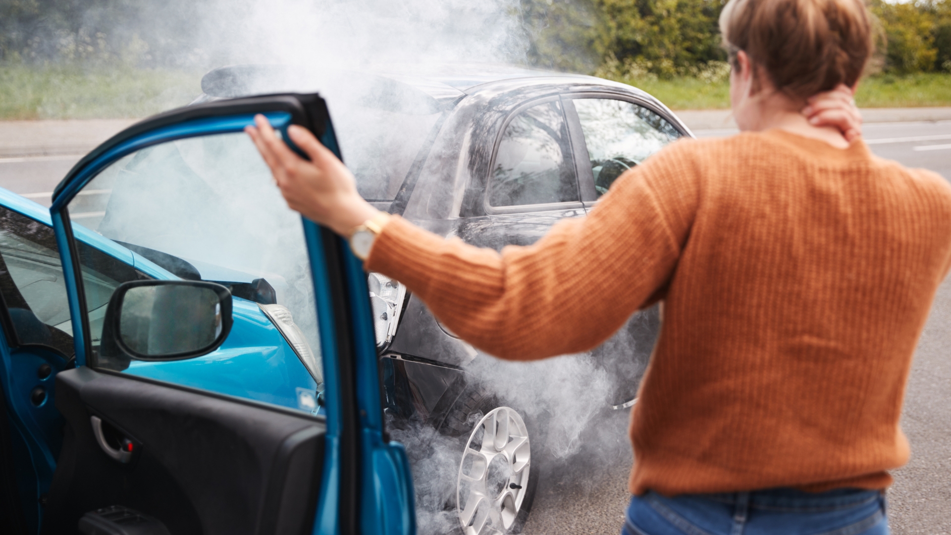 Two cars after a collision, with smoke rising and a person standing by an open car door holding their neck.