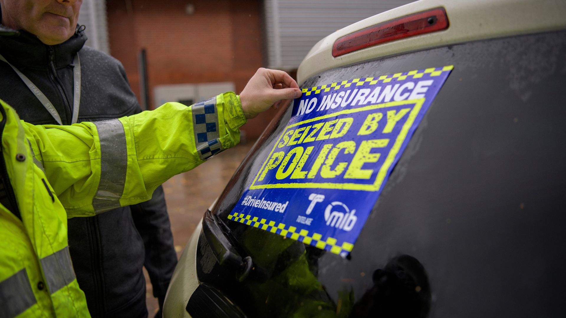 A high-visibility jacketed police officer placing a “No insurance seized by police” sticker on a car window.